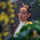 Mary is surrounded by plants while sporting a white turtleneck and two long, thin buns.